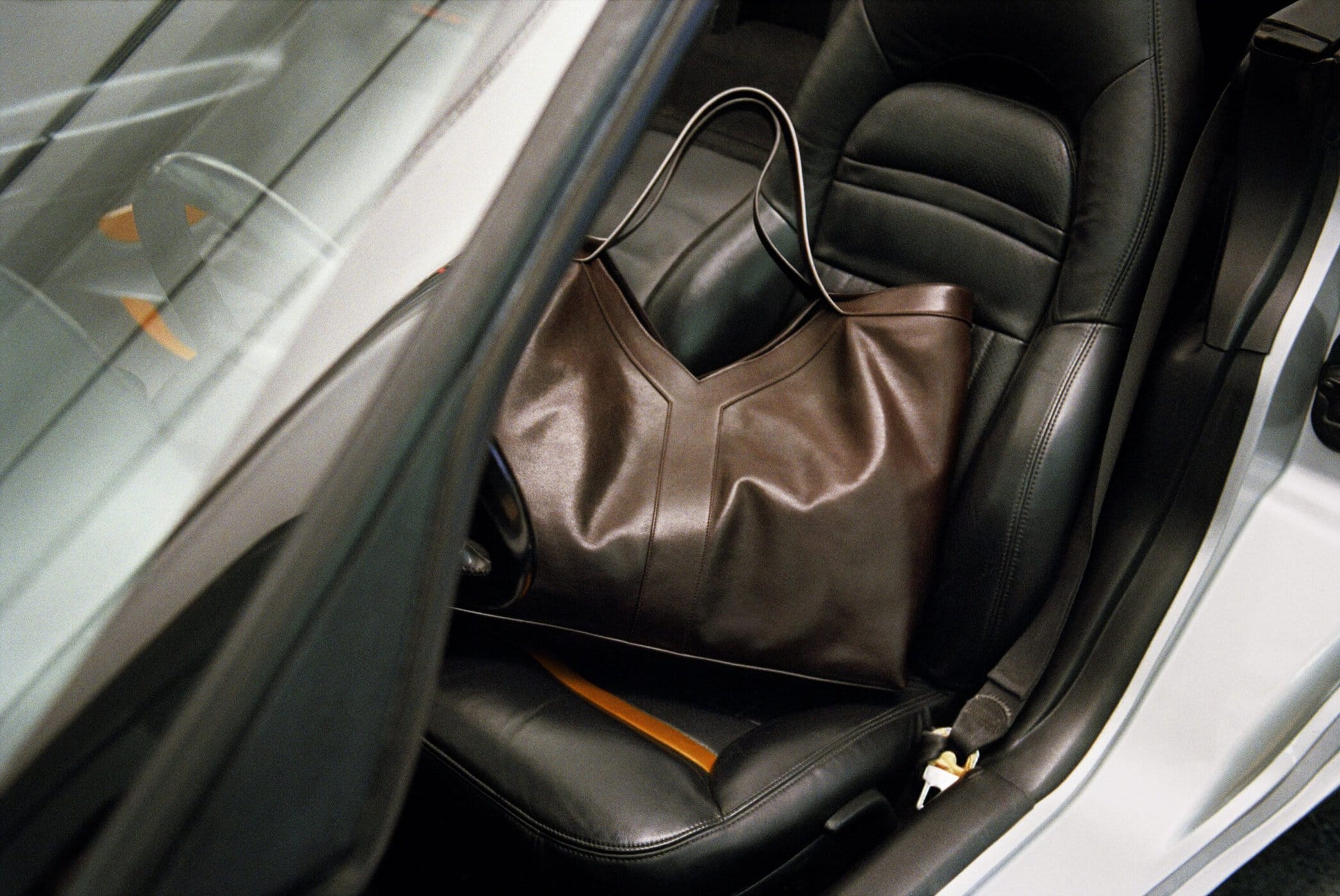 A dark brown leather tote bag resting on the black leather passenger seat of a convertible sports car.