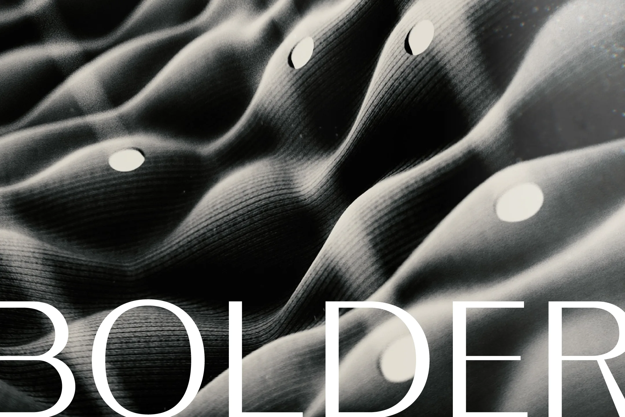 Monochrome close-up of undulating ribbed fabric with circular cutouts behind white typography reading BOLDER.