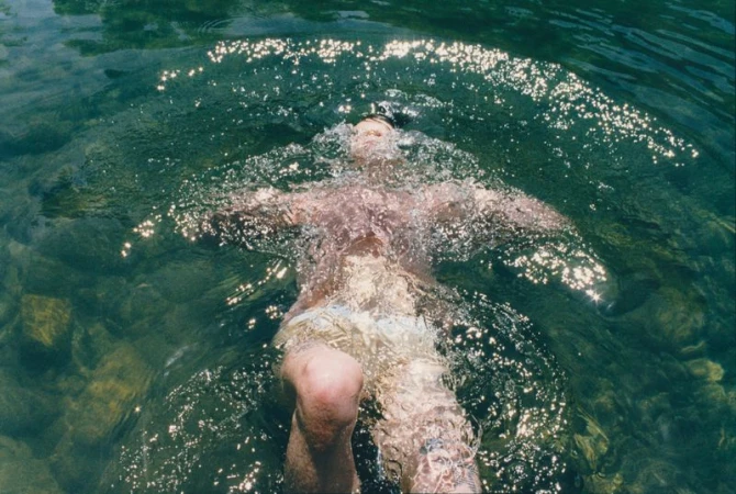 A man floating on his back just beneath clear green water with sun glinting off surface ripples.