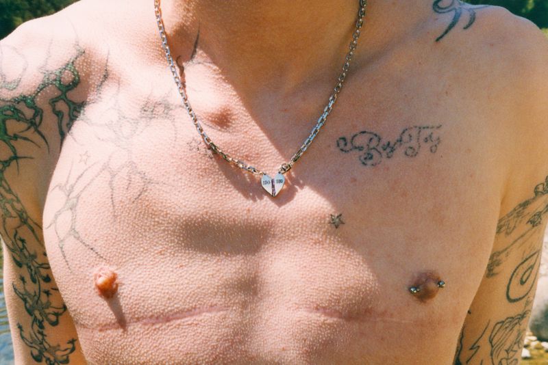 Close-up of Valentino Pavesi shirtless wearing a silver heart pendant necklace with visible chest tattoos and scars in sunlight.