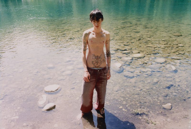 Model Valentino Pavesi standing shirtless with torso tattoos in clear shallow river water wearing brown jeans for Magliano Lele SS26.
