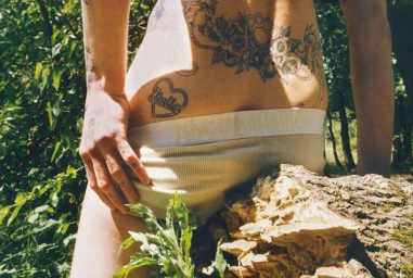 Close-up of a person's lower back with tattoos wearing Magliano ribbed underwear outdoors near a mossy tree trunk.