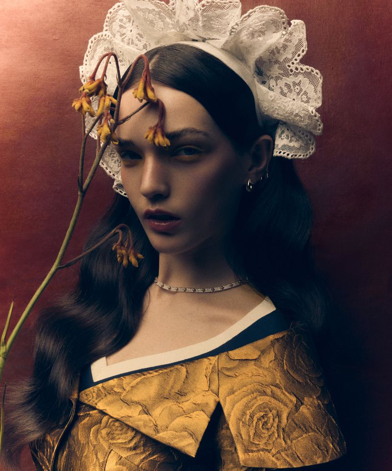 Portrait of a young woman with wavy hair wearing a lace bonnet and a golden floral dress, posing in a studio with a plant.