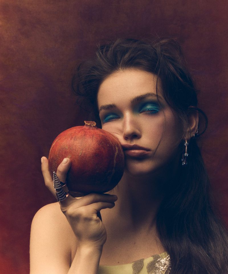 Brunette woman poses with a pomegranate in front of a warm brown background. She wears blue eyeshadow and a silver ring.