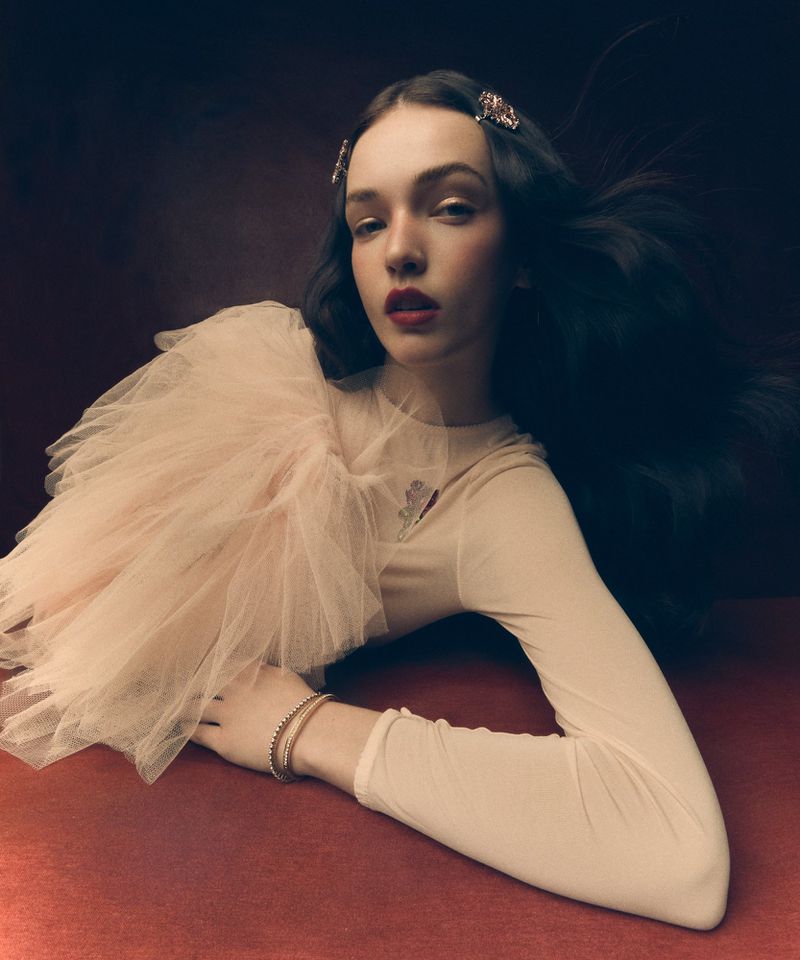 Portrait of a woman with long dark hair, wearing a pale pink long-sleeved top with a tulle shoulder detail, lying on a reddish-brown surface.
