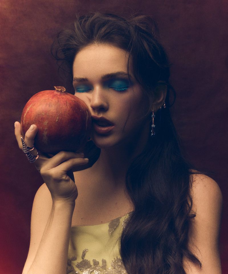 Brunette woman with closed eyes in soft lighting gently touches a vibrant pomegranate, wearing a yellow dress with ornate silver detail.