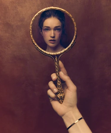 Elegant portrait of a young woman looking in a vintage, ornate hand mirror, held by a hand with a bracelet, on a bronze-colored background.