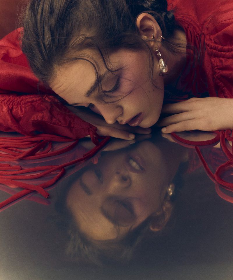 Close-up of a young woman lying down with her head resting on her hands. Her reflection is visible on the shiny surface below.