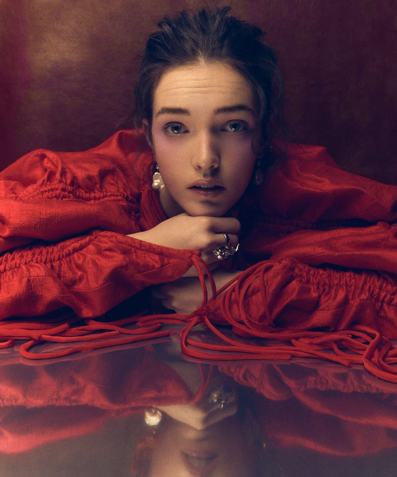 Close-up studio shot of a young woman with fair skin, wearing a red blouse with decorative drawstrings and pearl earrings.