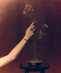 Close-up of a person's arm reaching out to touch delicate wildflowers arranged in a small metal bowl on a dark round table.