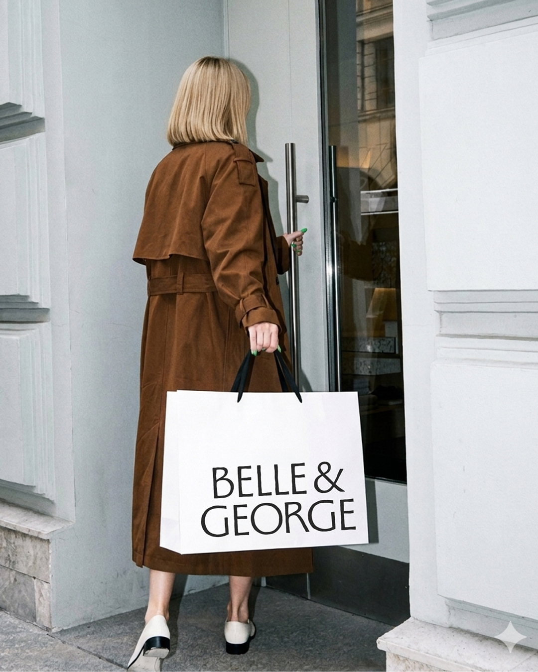 Rear view of a blonde woman in a brown trench coat holding a large white Belle & George shopping bag while opening a glass door.