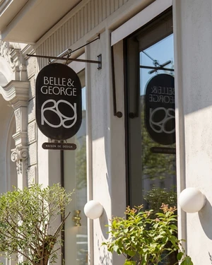 Black oval hanging storefront sign with white lettering for Belle & George mounted on a light-coloured architectural facade.