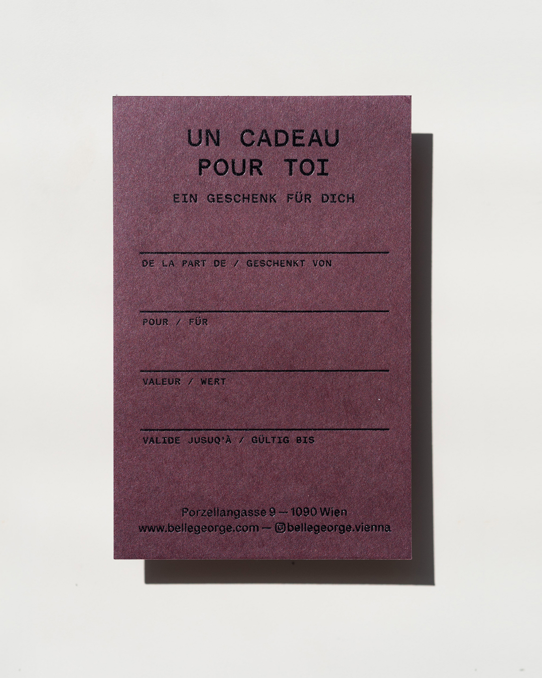 Vertical plum purple gift card with black French and German typography casting a sharp shadow on a white background.