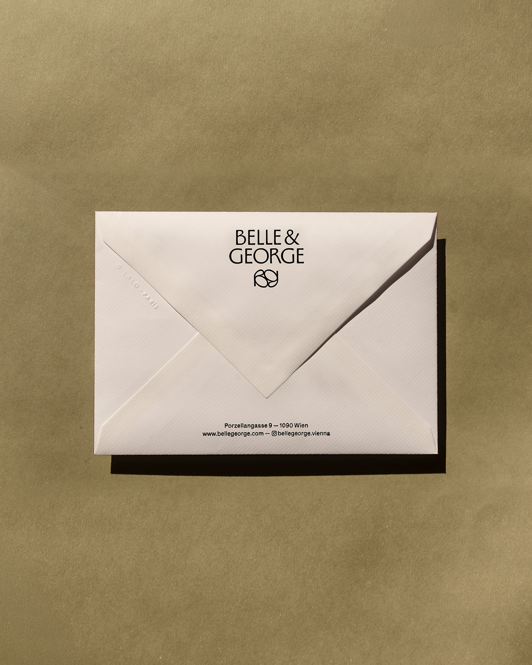 White paper envelope featuring Belle & George printed branding and monogram, placed on a smooth olive paper background.