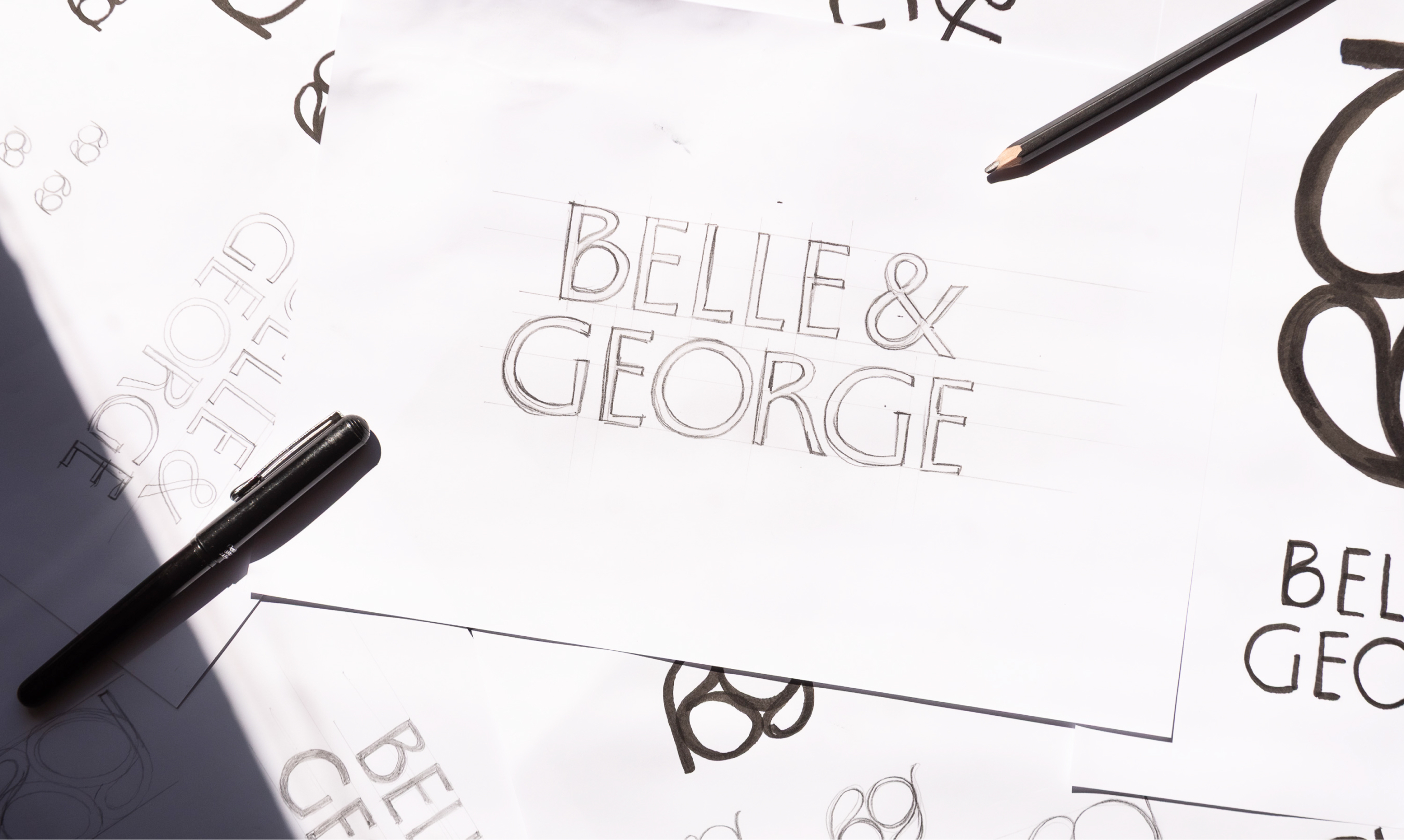 Hand-drawn pencil and ink typographic sketches of the Belle & George logo on white paper sheets with drawing pens.