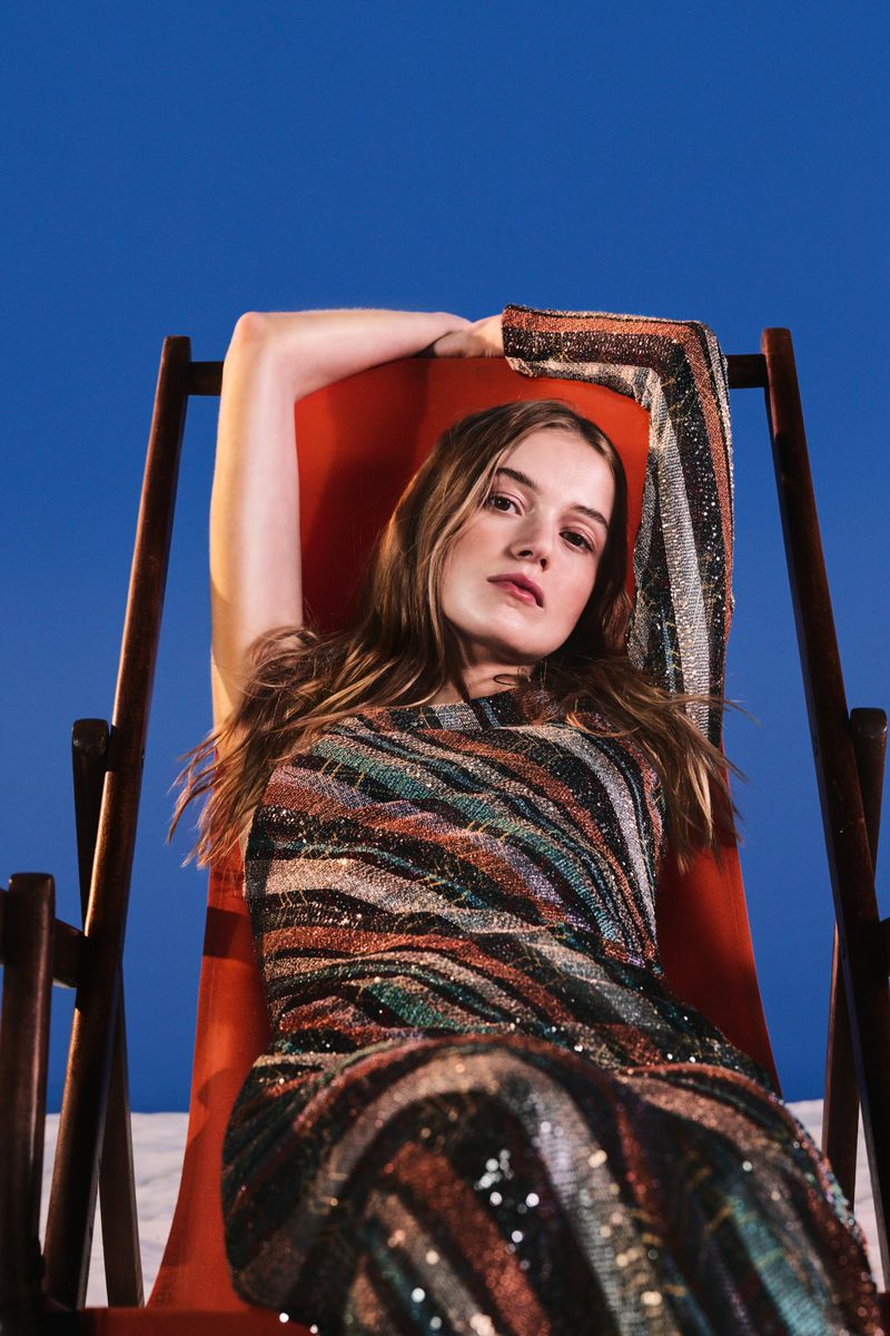 A woman reclining in an orange deck chair wearing a shimmering striped knit dress against a solid blue background.