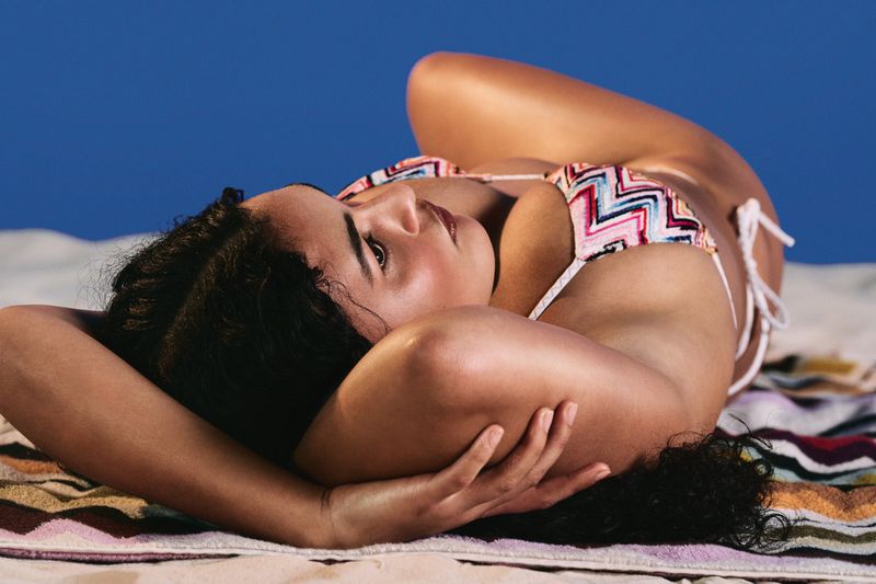 Woman reclining on a striped towel on the sand wearing a chevron patterned bikini top against a blue background.