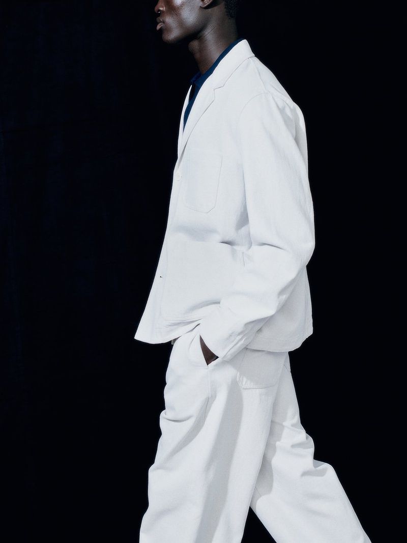 Model Gabriel Nhial wearing a relaxed off-white tailored jacket and matching trousers against a stark black studio backdrop.