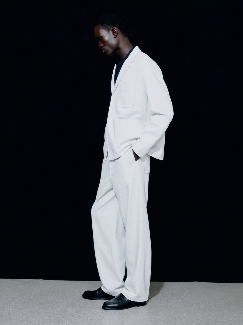 Model Gabriel Nhial stands in profile wearing a relaxed white suit and black loafers against a solid black studio backdrop.