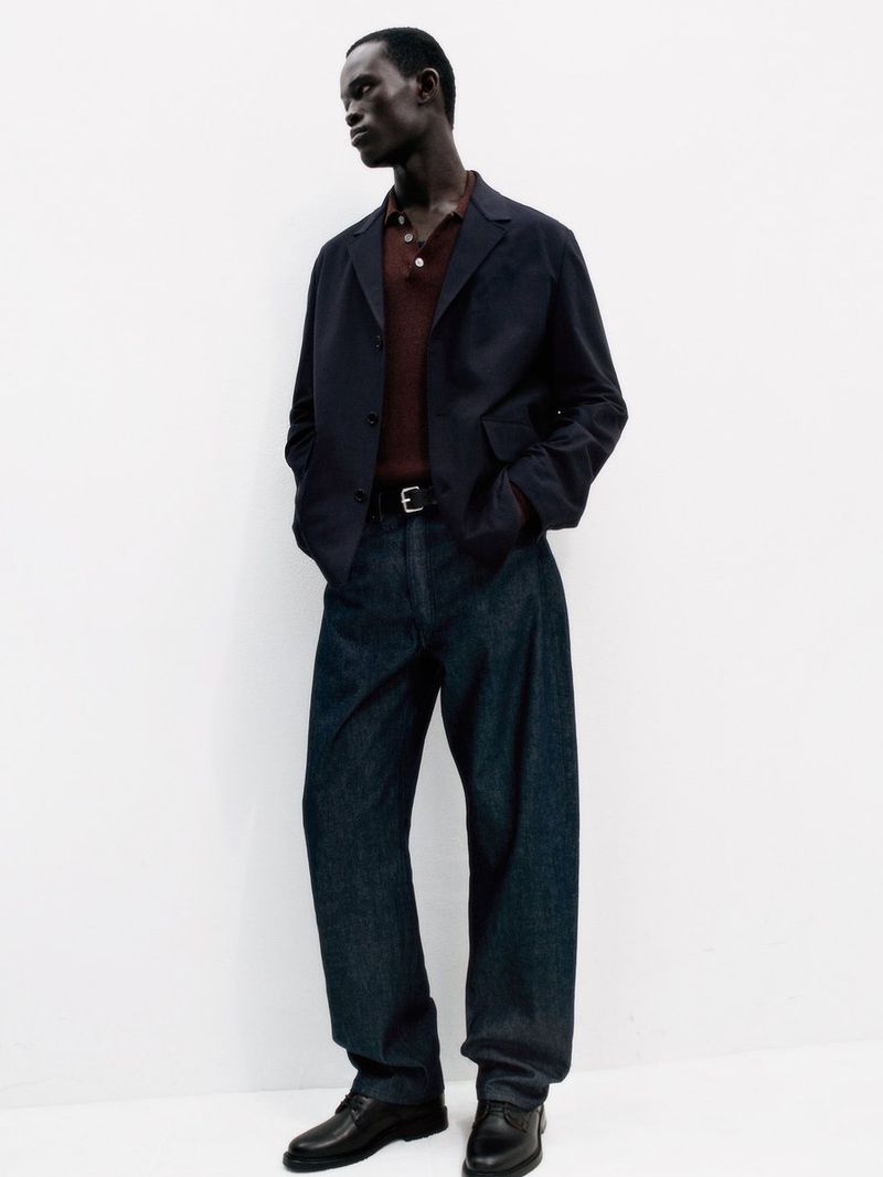 Model wearing dark blazer, brown polo shirt, and dark trousers against a plain white background