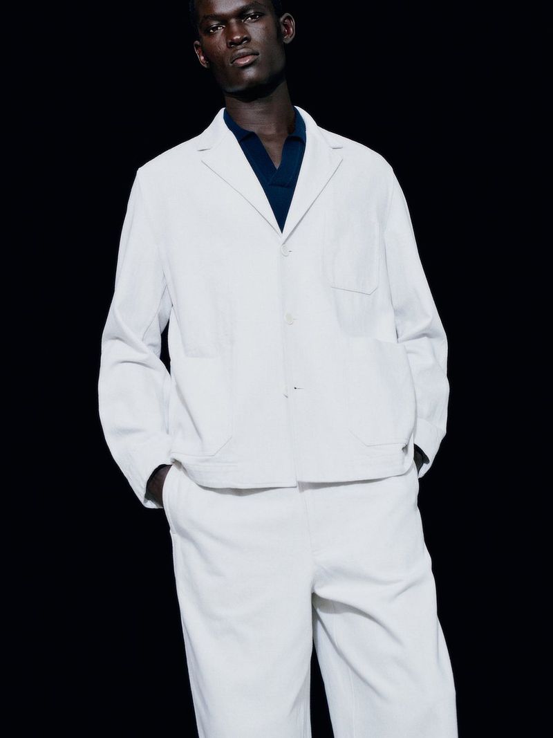 Model Gabriel Nhial wearing a white lightweight jacket, matching trousers, and dark collared shirt against a black background.