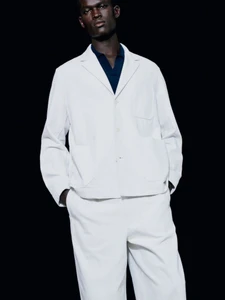 Model Gabriel Nhial wearing a white lightweight jacket, matching trousers, and dark collared shirt against a black background.