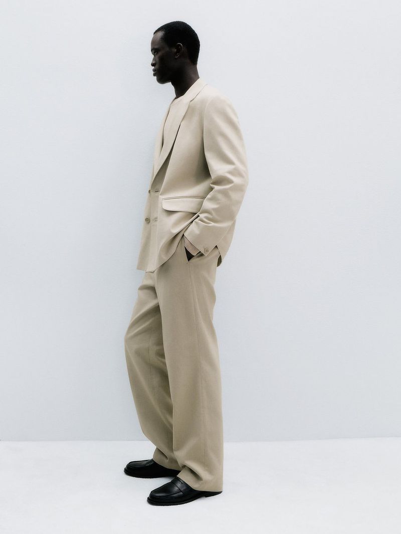 Gabriel Nhial posing in profile wearing a tailored beige suit and black loafers against a white studio background.