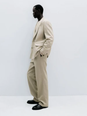 Gabriel Nhial posing in profile wearing a tailored beige suit and black loafers against a white studio background.