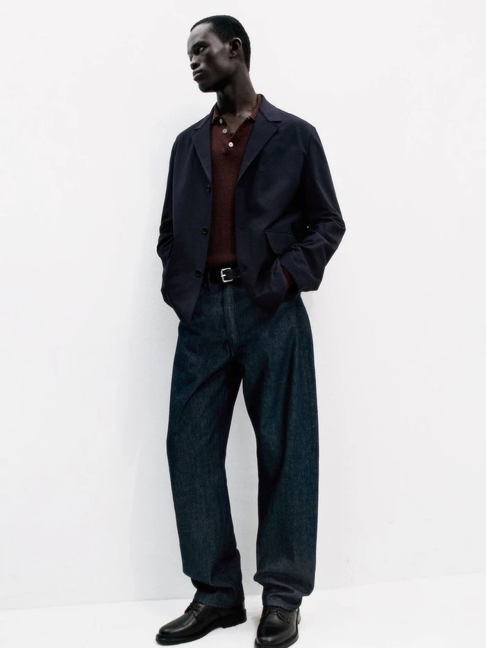 Model wearing dark blazer, brown polo shirt, and dark trousers against a plain white background