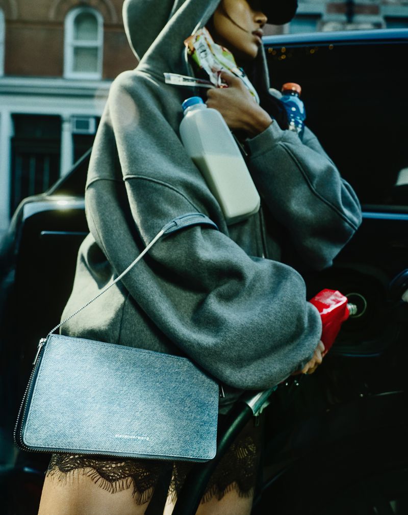 Model wearing an oversized grey hoodie holding a milk jug and handbag while holding a red gas pump nozzle.