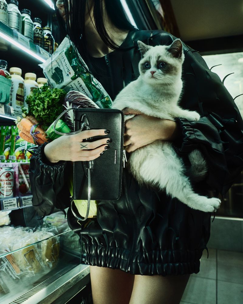 Model in a black jacket holding a cat, groceries, and an Alexander Wang black leather bag in a grocery store aisle.