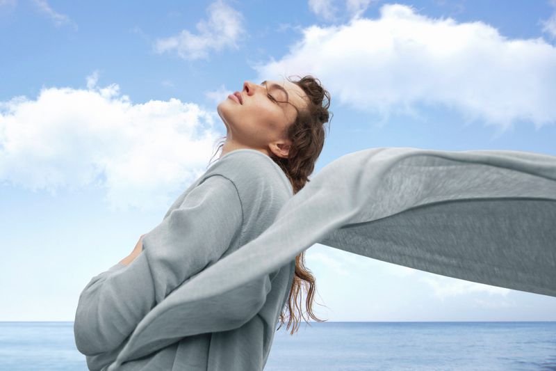Model Valerie Pasker with closed eyes tilting head up wearing light grey knitwear with fabric billowing against an ocean and sky background.