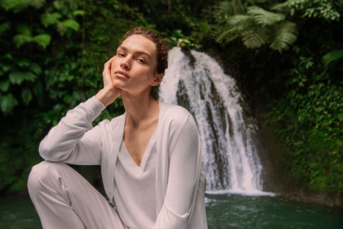 Model posing in an all-white knit cardigan and trousers against a lush tropical waterfall and foliage.