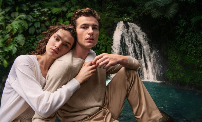 Man and woman modeling neutral knitwear embracing outdoors in front of a waterfall and lush foliage.