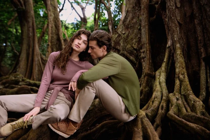 Models Valerie Pasker and Yulef Bopp seated amidst large tree roots wearing knitwear in a forest setting.