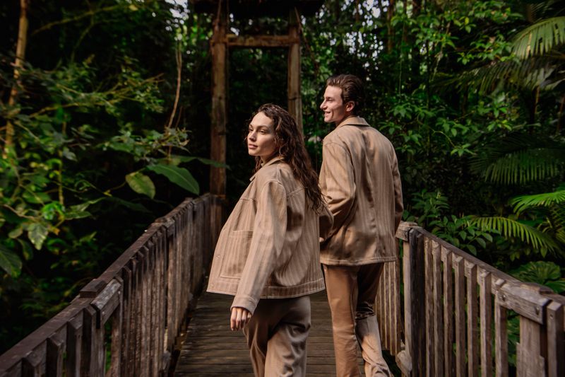 A woman and a man in matching beige jackets walking across a wooden bridge in a dense forest for the Falconeri S/S 26 campaign.