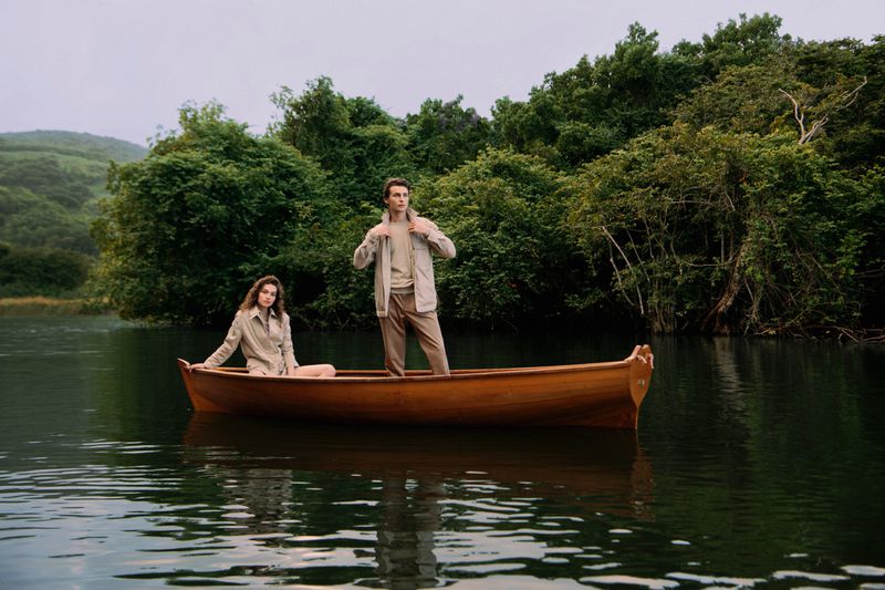 Two models in beige outerwear posing inside a wooden rowboat on a calm lake surrounded by lush green trees.