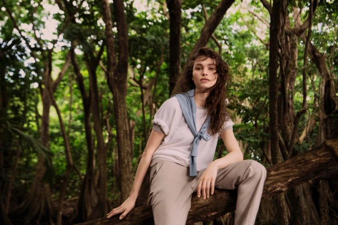 Model Valerie Pasker wearing a t-shirt, beige trousers, and a blue tied sweater, sitting on a tree branch in a dense green forest.
