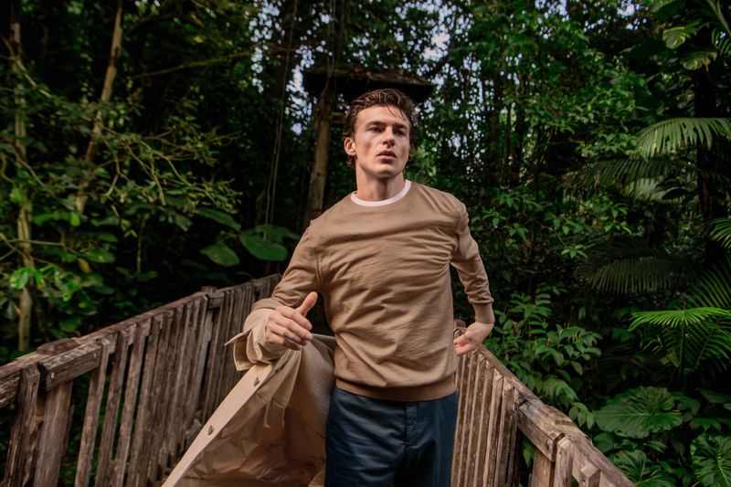 Model Yulef Bopp in a camel knit sweater and dark trousers walking across a wooden bridge in a lush tropical forest.