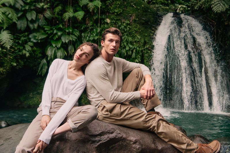 Man and woman sitting back-to-back on a large rock in front of a waterfall, wearing light knit sweaters and khaki trousers.