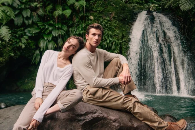 Man and woman sitting back-to-back on a large rock in front of a waterfall, wearing light knit sweaters and khaki trousers.