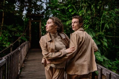 Two models wearing beige knit outerwear standing on a wooden bridge surrounded by lush green rainforest trees.