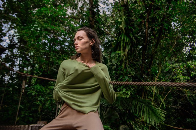Model Valerie Pasker wearing a green long-sleeve knit sweater and beige trousers against dense green forest foliage.