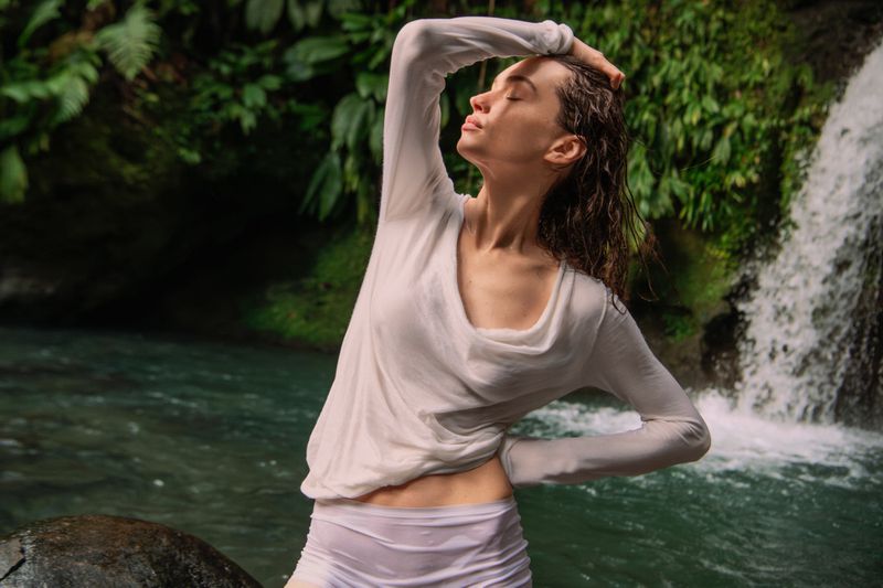 Model Valerie Pasker posing in a sheer white long-sleeve top in front of a tropical waterfall for Falconeri.