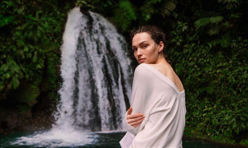 Model Valerie Pasker wearing an off-white knit sweater looking over her shoulder in front of a cascading waterfall.