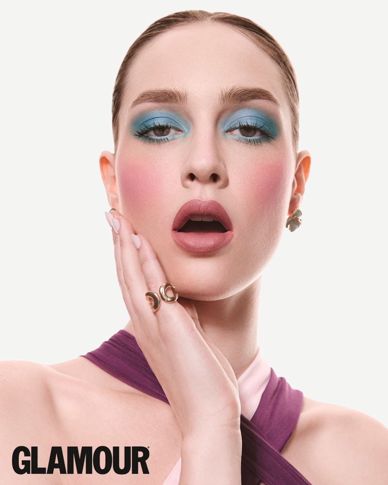 Woman with vibrant blue eyeshadow and pink blush posing with hand on cheek against a white background for Glamour.