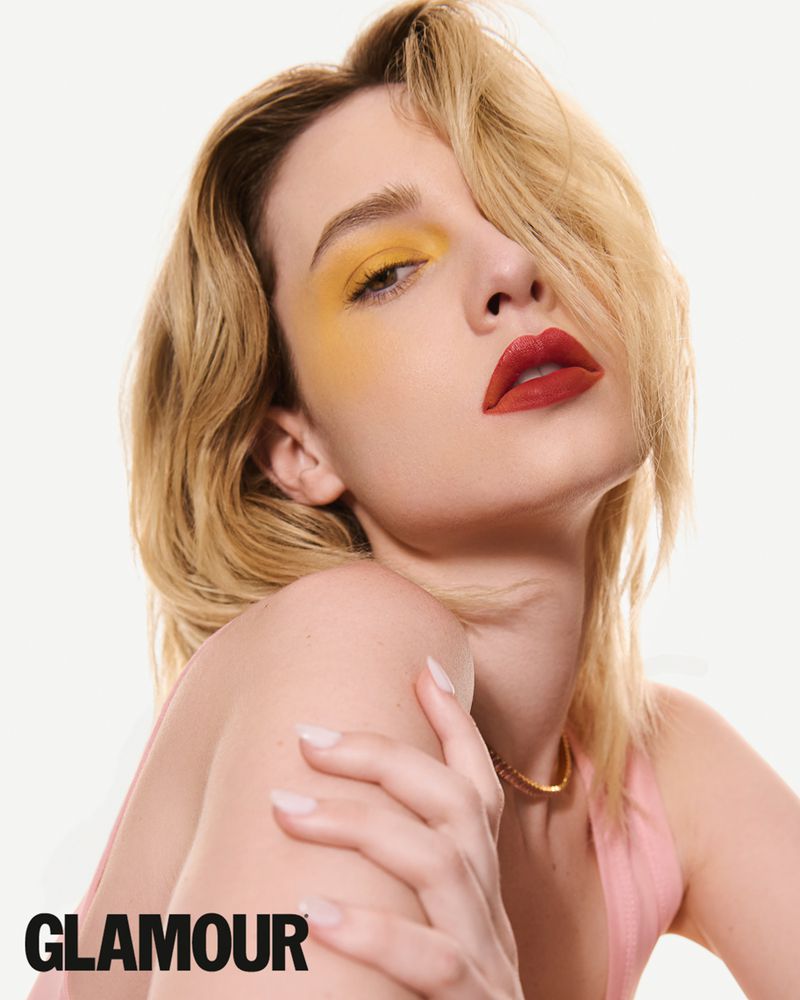 Close-up beauty portrait of a blonde woman wearing bold yellow eyeshadow and matte red lipstick against a white background.