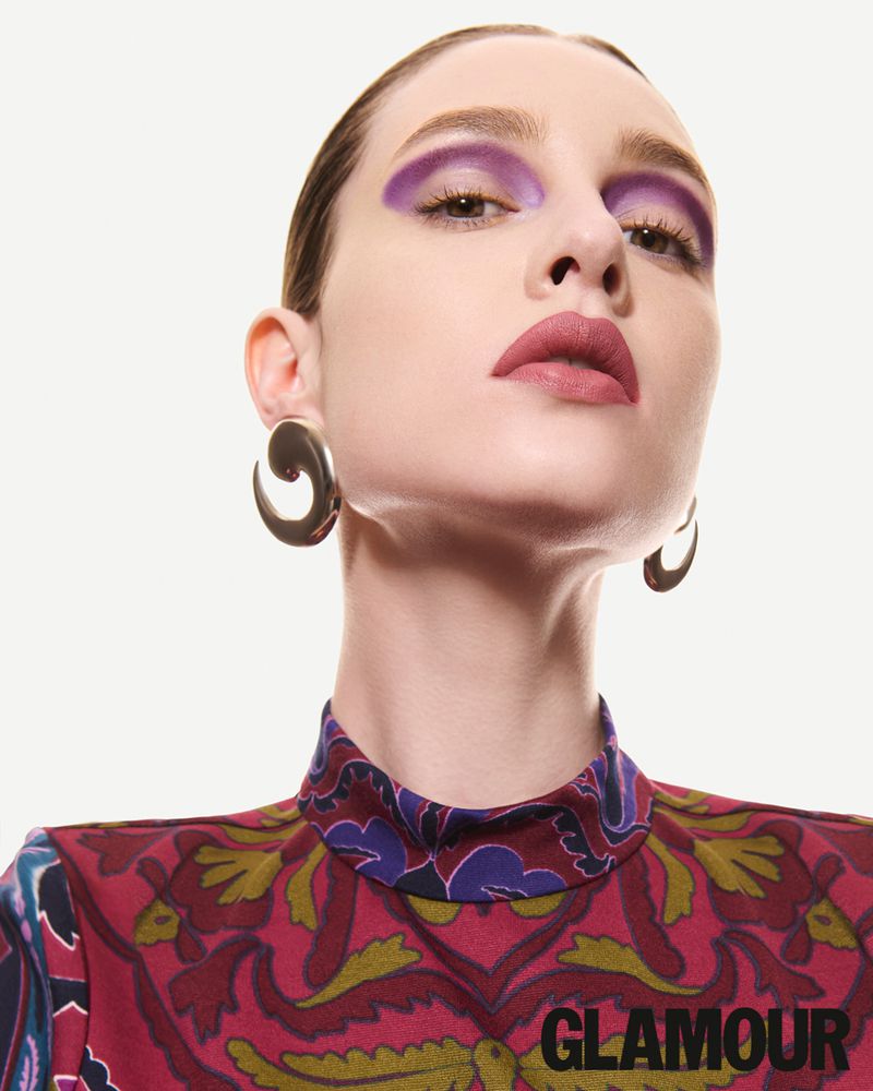 Close-up portrait of a woman with dramatic purple eyeshadow, sculptural earrings, and an ornate high-neck top against a white backdrop.