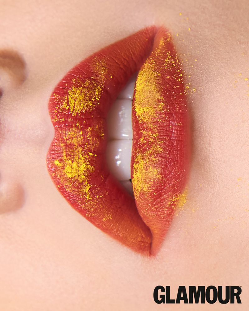 Extreme close-up of slightly parted lips with matte orange-red lipstick dusted with fine golden yellow powder.