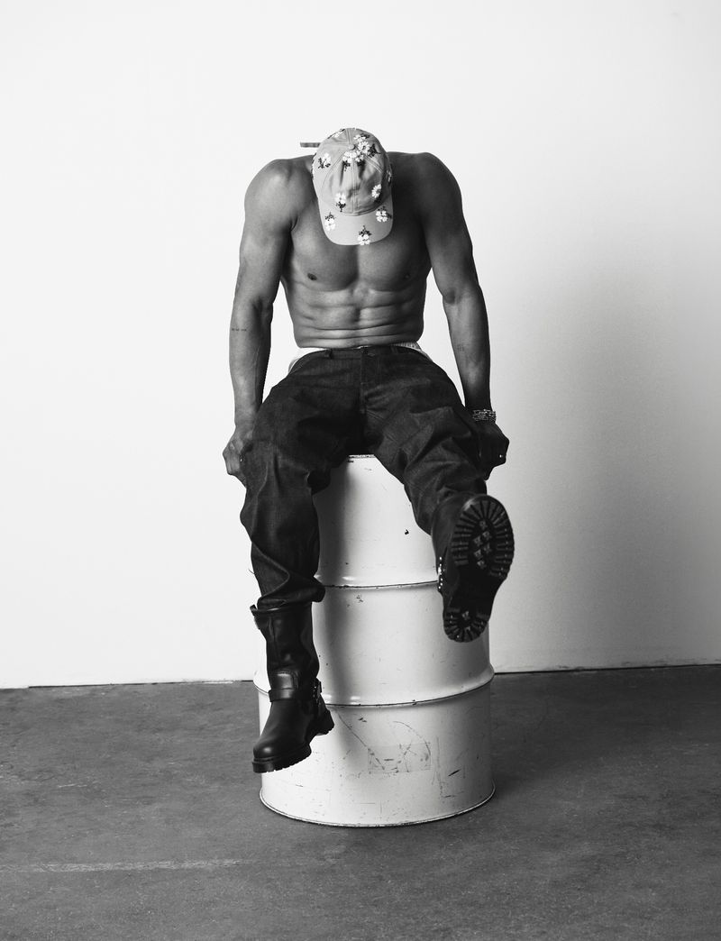 Shirtless man in dark jeans, boots, and a floral cap looking down while seated on a white metal drum against a plain studio background.